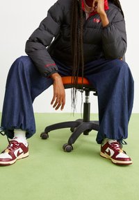 Zwarte pufferjack met rode accenten, donkergroene wijde denim broek die bij de enkels is aangetrokken, en bordeauxrode en witte Nike-sneakers op een groen tapijt.