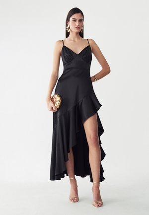 Frau in einem schwarzen asymmetrischen Kleid mit dünnen Trägern, goldenen Ohrringen, Armband, Clutch und hochhackigen Sandalen vor weißem Hintergrund.