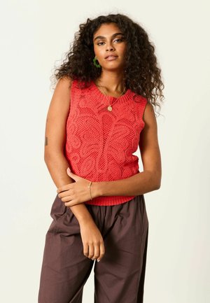 Woman with curly hair wearing a red sleeveless knit top, brown pants, green earrings, and a gold necklace and bracelet, posing against light background.