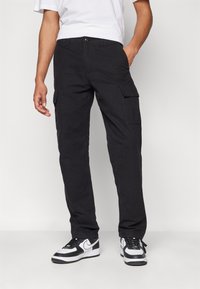 Černé pracovní kalhoty s několika bočními kapsami, rovný střih a odolný materiál z bavlněné směsi. Nošeny s bílými a černými teniskami.