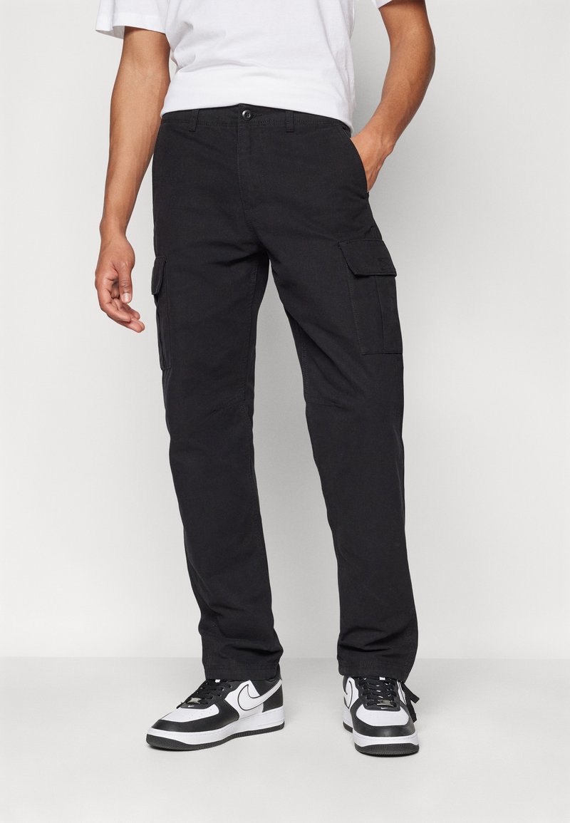 Černé pracovní kalhoty s několika bočními kapsami, rovný střih a odolný materiál z bavlněné směsi. Nošeny s bílými a černými teniskami.