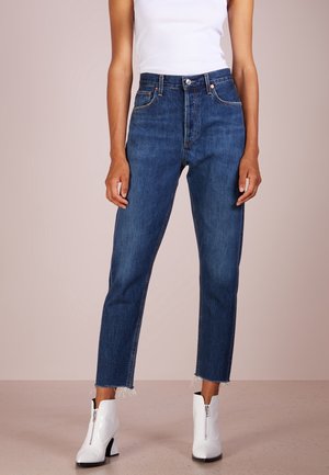 Osoba ubrana w ciemnoniebieskie jeansy z wysokim stanem i surowym wykończeniem, biały top bez rękawów oraz białe botki z przodu na zamek, stojąca na beżowej podłodze.