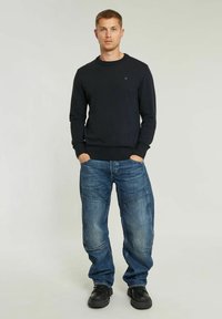 Felpa nera a collo rotondo in maglia con polsini a costine, abbinata a jeans in denim blu a vestibilità comoda con molteplici tasche e una finitura sbiadita.