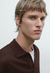 Hnědý polo tričko se strukturovaným límečkem, které je vrstveno přes bílé tričko s kulatým výstřihem. Detailní záběr muže s krátkými, texturovanými vlasy a světlou pletí.