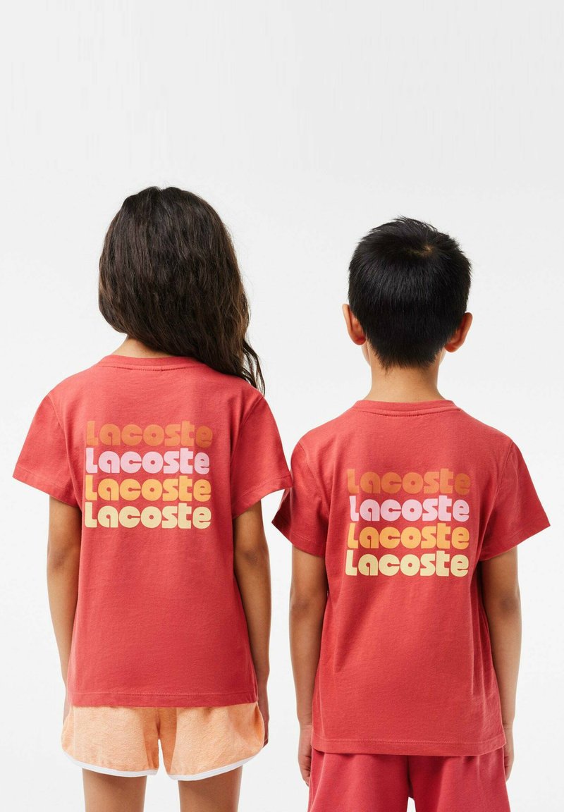 Rote Baumwoll-T-Shirts, die von hinten präsentiert werden, mit auffälligem "Lacoste"-Schriftzug in verschiedenen Farbtönen. Kurze Ärmel, lockere Passform, einfarbige Textur.