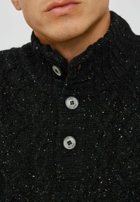 Schwarzer Strickpullover mit hohem Kragen, ausgestattet mit silbernen Knopfverschlüssen und einem strukturierten Muster aus unterschiedlichen Stichen.