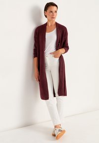 Lång vinröd cardigan i mjukt, texturerat material, bärs över en vit tanktop och vita jeans, med en enkel knappstängning. Avslappnade sneakers.