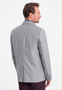 Vaaleanharmaa bleiseri, jossa on muotonsa mukaan räätälöity istuvuus, läpällä varustetut kaulukset ja yksi halkio, yhdistettynä tummiin housuihin. Pintakuvioitu kangas ja napitettu hihansuu.