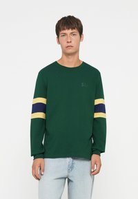 Green long-sleeve t-shirt made of cotton, featuring yellow and blue horizontal stripes on sleeves, with logo printed subtly on chest.