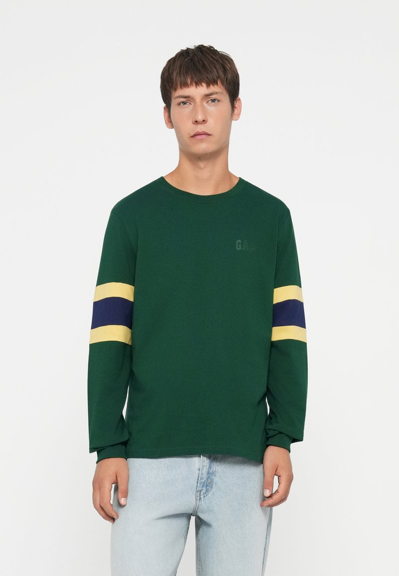 Green long-sleeve t-shirt made of cotton, featuring yellow and blue horizontal stripes on sleeves, with logo printed subtly on chest.