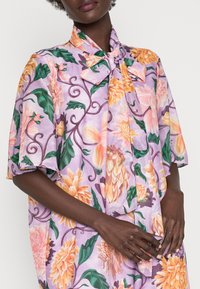 Camisa floral en lavanda con grandes flores naranjas y rosas, hojas verdes y un escote con lazo; ajuste holgado con mangas cortas.