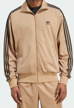 Mężczyzna w beżowej kurtce dresowej i spodniach Adidas z czarnymi paskami na rękawach, stojący na neutralnym tle.