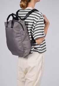 Mochila de nailon gris con textura acolchada, doble asa superior, correas negras ajustables y un diseño minimalista. Camiseta a rayas y pantalones claros puestos.