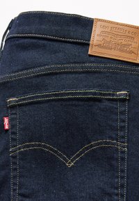 Calças de ganga de denim azul-escuro com um remendo de couro castanho com marcação em relevo, costura visível e uma pequena etiqueta de logo vermelho de lado.