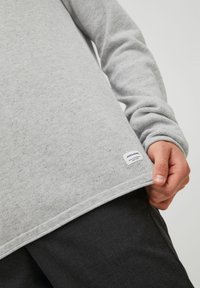 Osoba ubrana w jasnoszary, teksturowany, długi rękaw, z małą białą metką marki blisko dołu, oraz ciemnoszare spodnie.