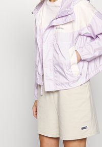 Lichtpaarse windjack met elastische manchetten, voorzakken en een verstelbare zoom. Gepaard met beige shorts met een zijlabel.