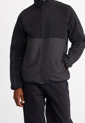 Veste polaire noire zippée avec partie supérieure et manches texturées, panneau avant inférieur lisse, poches latérales, portée avec un pantalon noir.