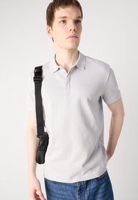 Polo shirt gris clair à manches courtes, avec un petit col, des détails de couture verticaux et un logo. Associé à un jean bleu et un sac à épaule noir.