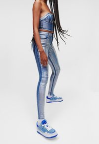 Denim-kaksiosainen asu, jossa on olkaimeton cropped-top ja korkeavyötäröiset kapeat farkut haalistuneessa sinisessä, kiiltävällä pinnalla, yhdistettynä sinisiin lenkkareihin.