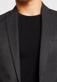 Blazer noir à rayures fines en tissu texturé, avec un t-shirt noir à col rond en dessous, mettant en valeur les revers et des détails de couture subtils.
