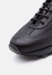 Baskets en cuir noir avec couture détaillée, bout rond et semelle épaisse texturée sur un fond blanc.