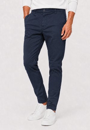 Man draagt een slim-fit marineblauwe broek, witte sneakers en een wit shirt met lange mouwen, staat met handen gedeeltelijk in de zakken.