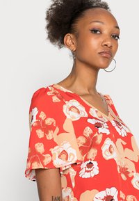 Blusa floral vermelha com mangas curtas e evasê, apresentando flores em creme e laranja, decote em V e um tecido leve e texturizado.