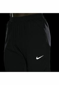 Nahaufnahme einer Person, die schwarze Sporthosen mit elastischem Bund und einem weißen Nike-Swoosh-Logo auf dem Oberschenkel trägt.