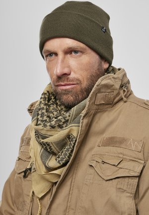 Bărbat cu barbă deschisă la culoare, purtând o căciulă verde olive, o jachetă tactică bej și un fular bej și negru cu model, pe un fundal simplu.