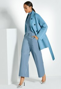 Helles blaues Doppelreiher-Mantel über einem passenden Rollkragenpullover und weitem Hosen. Metallic-silberne High-Heels runden das Outfit ab.