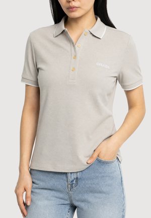 Femme portant un polo beige clair à manches courtes avec boutons et logo Gisada brodé, associé à un jean bleu clair, main dans la poche.