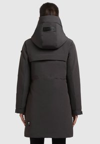 Parka nera, impermeabile, con colletto alto e cappuccio regolabile. Presenta un pannello posteriore e dettagli con logo. Texture liscia e opaca, design che arriva fino al ginocchio.