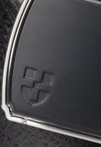 Czarna skórzana metka z srebrną ramką, z wytłoczonym wzorem logo, który zawiera kratki oraz półokrągły kształt na dole.