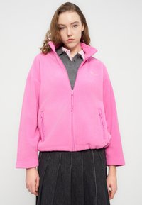 Ροζ φλις μπουφάν με ψηλό γιακά, φερμουάρ μπροστά και δύο πλαϊνές τσέπες με φερμουάρ. Φορεμένο πάνω από γκρι πουκάμισο και μαύρη πλισέ φούστα.