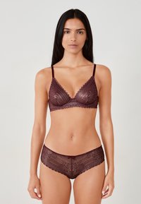 Donna che indossa un set coordinato di reggiseno in pizzo bordeaux e culotte a vita alta, in piedi contro uno sfondo chiaro e uniforme.