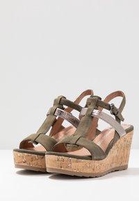 Sandales à talon compensé vert olive avec une plateforme en liège, présentant des accents métalliques argentés et un design ouvert à l'avant. Sangle ajustable avec détail de boucle.