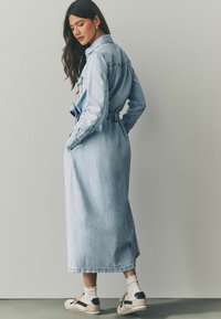 Jeans trench coat i ljusblå med bälte i midjan, långa ärmar och sidofickor. Matchad med beige sneakers och vita strumpor.
