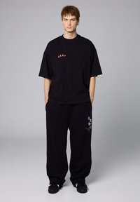 Czarny, oversized t-shirt z czerwonym napisem "GRMY" na piersi, w połączeniu z szerokimi czarnymi spodniami z niebieskimi i czerwonymi grafikami. Gładka tekstura materiału.