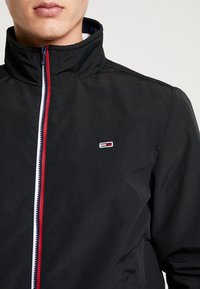 Czarna kurtka z wysokim kołnierzem, zamkiem na całej długości z czerwonym, białym i niebieskim akcentem oraz małym haftowanym logo na piersi.