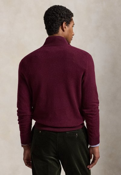 Mies, jolla on päällään istuva viininpunainen pitkähihaisen neule ja tummanvihreät corduroy-housut, seisoo selin katsojaan.