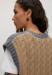 Gebreide vest met een tan kabelontwerp en een grijze ribbelkraag. De stof lijkt gestructureerd en er wordt een wit T-shirt onder gedragen.