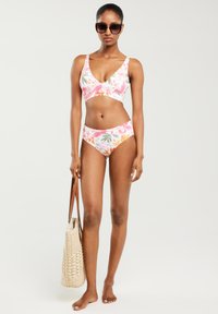 Kostium kąpielowy w kwiatowy wzór w odcieniach różu, bieli i zieleni. Górna część ma dekolt w kształcie litery V, a do niej dopasowane majtki o średnim stanie. Pleciona torba.