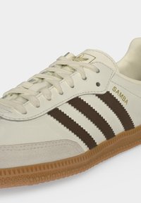 Κοντινή λήψη μπεζ αθλητικού παπουτσιού Adidas Samba με καφέ ρίγες, λαστιχένια σόλα, λευκά κορδόνια και χρυσό λογότυπο στο πλάι και στη γλώσσα.