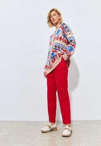 Neulecardigan vaaleanpunaisena, jossa on sinisiä, punaisia ja mustia kuvioita, yhdistettyinä punaisiin housuihin ja valkoisiin kenkiin, joissa on beige aksentti. Sujuva rakenne.