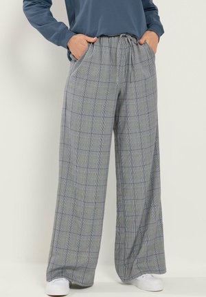 Person trägt weite, graue karierte Hose mit Kordelzugbund, Hände in den Taschen, kombiniert mit weißen Sneakers und einem blauen Oberteil.