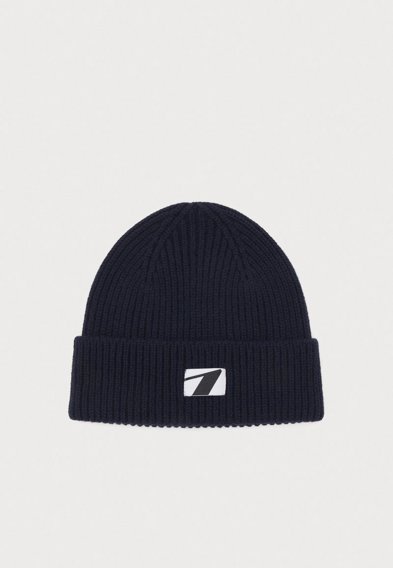 Bonnet côtelé bleu marine avec revers et une étiquette blanche rectangulaire présentant un logo stylisé du chiffre sept en noir.