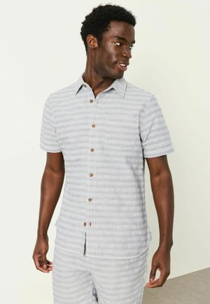 Mann trägt ein hellgraues, kurzärmeliges Hemd mit Knöpfen und passende Hose mit dezenten vertikalen Streifen, schaut nach rechts und lächelt.