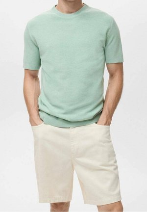 Homme portant un pull vert clair à manches courtes et un short couleur crème, mains dans les poches, sur un fond uni.