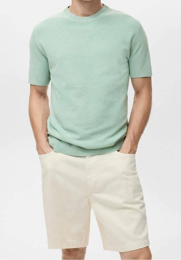 Homme portant un pull vert clair à manches courtes et un short couleur crème, mains dans les poches, sur un fond uni.