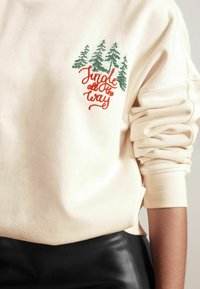 Sweat-shirt couleur crème avec des arbres verts brodés et le texte en cursive rouge "Jingle all the way." Manches retroussées et texture de tissu doux.